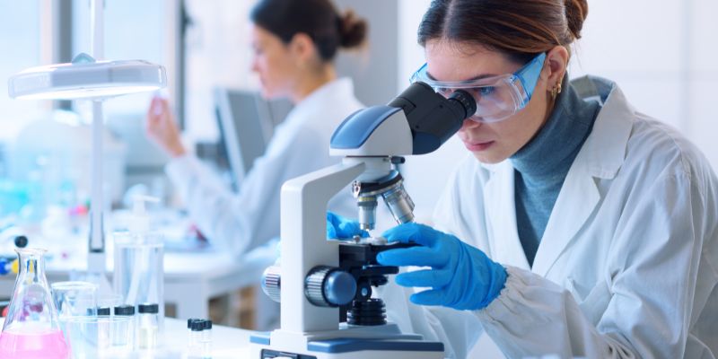 Woman looking through a microscope with another person in the background. They are in a laboratory.
